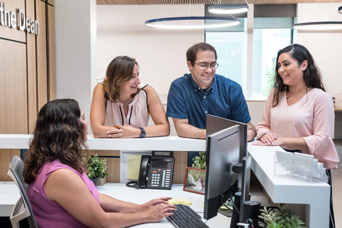 Three people stand and one person is sitting behind a desk