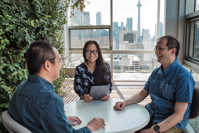 Three people sit at a round table with a green leafy wall to the side