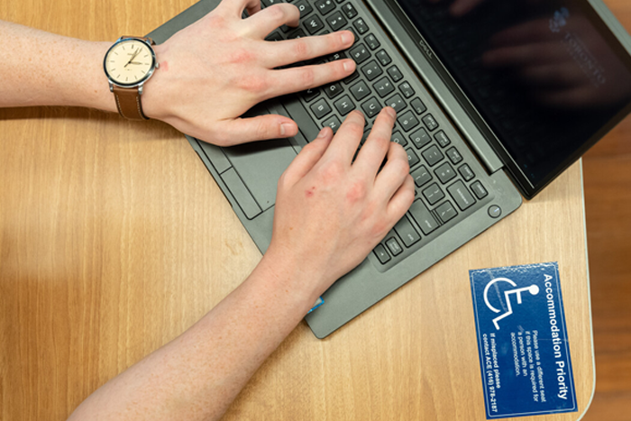 A person types on a laptop and there is an accommodations priority sticker on the desk