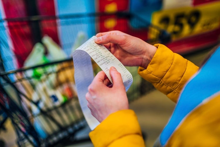 Woman looks at a grocery shopping receipt with a full shopping cart in the background