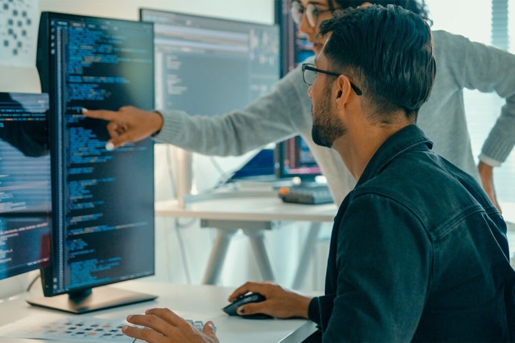 a male and female worker look over computer code in front of a computer monitor 