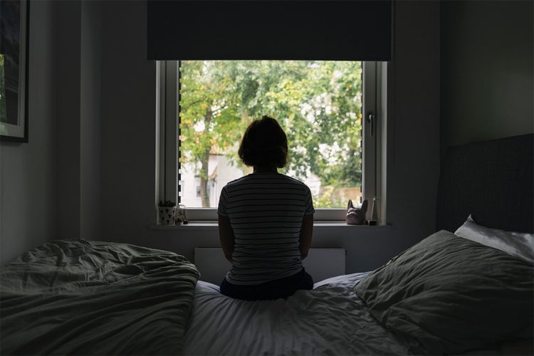 an anonymous woman in her 40s sits on her bed looking out the window