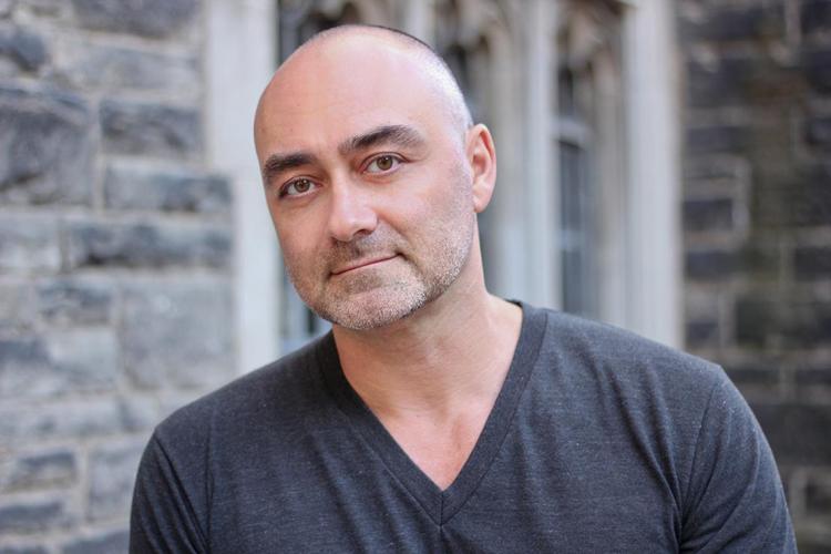 John Paul Ricco stands in front of a brick wall, wearing a t-shirt