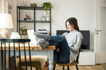 young woman with feet up on desk smiling while talking to someone on her laptop