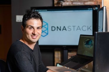 Marc Fiume sits at a computer, a monitor displaying the DNAstack logo is in the backbround