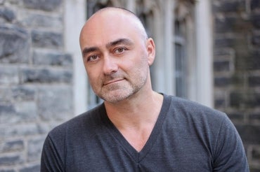 John Paul Ricco stands in front of a brick wall, wearing a t-shirt