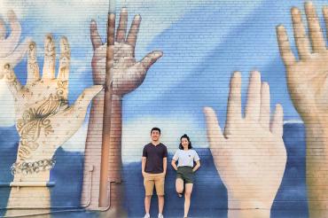 Weiwei Li and Catherine Chan stnad in front of a large mural on a wall depicting hands of different colours raised up