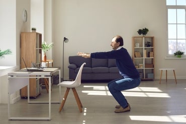 a man does a squat with arms extended behind his home office desk chair