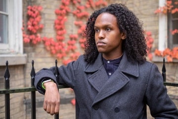A photo of Egag Egag wearing a wool top coat leaning on a wrought iron fence with red ivy in the background